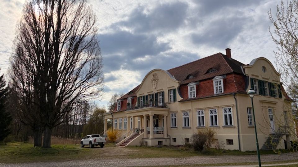 Großes, zweigeschossiges Herrenhaus mit hellem Putz, rotem Mansarddach und mehreren Gaubenfenstern, davor eine Treppe zu einem überdachten Eingang. Das Gebäude steht in einer offenen Grünanlage mit hohen, unbelaubten Bäumen, einem Kiesweg und bewölktem Himmel.