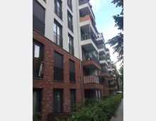 Mehrstöckiges modernes Wohngebäude mit einer Fassade aus rotem Backstein und weißem Putz. Die Wohnungen verfügen über Balkone mit Geländern, einige dekoriert mit Pflanzen oder Wäsche, und vor dem Gebäude befindet sich eine Hecke entlang eines gepflasterten Gehwegs.