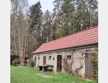Ein langgestrecktes Gebäude mit hellem Putz und rotem Ziegeldach, ausgestattet mit kleinen Fenstern und Holztüren. Im Vordergrund eine Holzbank und ein Tisch, umgeben von einer Wiese; im Hintergrund dichter Wald und kahle Bäume.