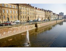 Eine Uferpromenade mit historischen Sandsteingebäuden aus mehreren Stockwerken, die parallel zu einem ruhig fließenden Kanal verläuft. Der Kanal wird durch eine niedrige Backsteinmauer begrenzt, und im Vordergrund ist ein kleiner Springbrunnen zu sehen.