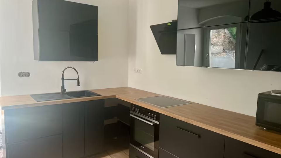 Moderne L-förmige Küche mit mattschwarzen Schränken und Holzarbeitsplatte, integriertem Spülbecken mit schwarzer Armatur sowie Herd und Backofen. An der Wand befinden sich glänzende, schwarze Hängeschränke und eine schräg montierte Dunstabzugshaube; heller Holzboden und Deckenleuchte ergänzen den Raum.