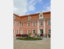 Rosa Gebäude mit symmetrischen weißen Fensterrahmen und Zierelementen, einer Dachuhr und roten Ziegeln. Im Vordergrund ein runder Blumenbeet-Bereich auf einem gepflasterten Hof, umgeben von parkenden Autos und stehenden Personen.