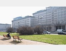 Weitläufiger Platz mit Grünflächen, Bänken und Büschen im Vordergrund, auf denen eine Person sitzt, während im Hintergrund große symmetrische Wohn- und Bürogebäude im monumentalen Stil der Nachkriegszeit zu sehen sind. Verkehrsstraße mit parkenden und fahrenden Autos sowie Laternenmasten trennt die Gebäude von den Grünanlagen.