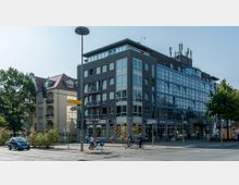 Moderne Straßenecke mit einem mehrstöckigen Gebäude in dunkler Fassade und großen Fensterfronten, neben einem älteren gelben Wohnhaus mit Dachgauben und Balkonen. Im Vordergrund eine Kreuzung mit Radfahrern, einem Auto und Verkehrsschildern.