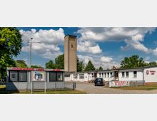 Feuerwache in Lübben mit einem langen, weißen Flachbau und einem hohen, rechteckigen Turm im Hintergrund. Im Vordergrund steht ein Werbeschild der Jugendfeuerwehr neben einem Parkplatz, umgeben von Bäumen und einer Wiese.
