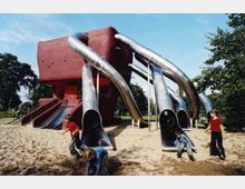 Ein moderner Spielplatz mit einem großen roten Gebäudekomplex und mehreren langen, silbernen Röhrenrutschen. Kinder spielen auf einem Sandboden umgeben von Bäumen und grüner Vegetation.