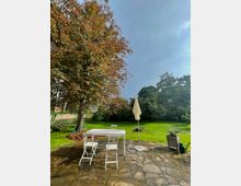 Ein Garten mit einer gepflasterten Terrasse, auf der weiße Gartenmöbel und ein geschlossener Sonnenschirm stehen. Im Hintergrund sind eine Rasenfläche, ein großer Baum mit herbstlich verfärbten Blättern und weitere Büsche und Bäume zu sehen.