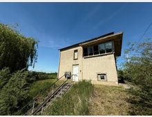 Zweistöckiges Gebäude mit beigem Putz und Flachdach, teils mit großen Fenstern in der oberen Etage. Eine Metalltreppe führt über eine grasbewachsene Böschung zum Eingang; im Hintergrund sind Bäume und blauer Himmel zu sehen.