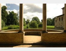 Steinveranda mit Rundbank und Säulen, die den Blick auf einen Park mit Bäumen, einer Wiese und einem kleinen Teich freigibt; rechts im Bild ist ein Teil eines historischen Gebäudes erkennbar.