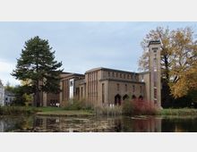 Backsteingebäude im Stil der frühen Moderne mit vertikalen Fensterlinien und einem hohen Turm, umgeben von Bäumen und einem Teich mit Wasserpflanzen im Vordergrund.