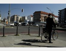 Straßenszene an einer Kreuzung mit mehreren Autos und Gebäuden im Hintergrund, darunter ein rotes Ziegeldachgebäude. Im Vordergrund steht eine Person mit einem Fahrrad auf einem Gehweg.