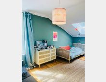 Ein gemütliches Schlafzimmer mit einer hellblau gestrichenen Wand, einem Tagesbett mit grauem Holzrahmen und blauen Bettbezügen. Links steht eine cremefarbene Kommode mit Rattanfronten, daneben lange hellblaue Vorhänge vor einem Fenster; der Raum ist mit Holzboden und einem Sisalteppich ausgestattet.