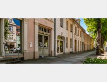 Straßenzug mit historischen, pastellfarbenen Gebäudefassaden mit hohen Fenstern und verzierten Türrahmen, teils mit Ladeneingängen im Erdgeschoss. Breiter Gehweg mit Kopfsteinpflaster und großen Straßenbäumen entlang der rechten Seite.