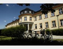 Gelbes, zweistöckiges Gebäude mit symmetrischen Fenstern und einem zentralen, runden Eingang mit Säulen. Im Vordergrund sind mehrere geparkte Fahrräder und Büsche zu sehen, der Himmel ist klar und blau.