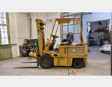 Ein gelber Gabelstapler steht in einer Werkstatt mit offenen Türen, Industriemaschinen und Werkzeugen im Hintergrund. Die Umgebung hat helle Wände, ein großes Fenster mit grünem Rahmen sowie teilweise sichtbare Lagergegenstände.