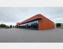 Backstein-Gebäude mit abgerundeten Ecken und einer Fassade aus großen Glasflächen, umgeben von einer gepflasterten Fläche, im Hintergrund Bäume und parkende Autos. Der Himmel ist bedeckt.