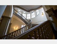 Eine geschwungene historische Treppe mit verzierten Balustraden in einem hellen Treppenhaus; große, bogenförmige Fenster lassen natürliches Licht herein, und an der Wand befinden sich dekorative Wandlampen.