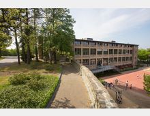 Ein großes Backsteingebäude mit mehreren Fenstern steht neben einem Schulhof mit Spielgeräten und spielenden Kindern. Links führt ein gepflasterter Weg entlang einer Steinmauer durch eine Grünanlage mit Bäumen und Sträuchern.