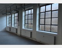 Innenraum eines industriellen Loft- oder Bürogebäudes mit weiß gestrichenen Ziegelwänden und großen, schwarz gerahmten Sprossenfenstern entlang der Wand. Unter den Fenstern sind mehrere Heizkörper angebracht; an der Decke verlaufen sichtbare Kabeltrassen und Leitungen.