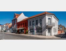 Straßenszene mit zweistöckigen Häusern im Altbaustil, teilweise in hellen und roten Farben gestrichen, mit Geschäften und Restaurants im Erdgeschoss. Der Gehsteig ist mit Straßenlaternen und Außensitzplätzen ausgestattet, während die Straße gepflastert und wenig befahren ist.