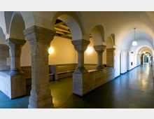 Ein langer Flur mit einer Reihe von Rundbögen, die auf massiven steinernen Säulen ruhen, zeigt eine Mischung aus historischer Architektur und moderner Innenbeleuchtung. Der Boden ist glatt und dunkelgrün, während die Wände hell gestrichen sind und durch angebrachte Wandlampen beleuchtet werden.