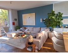 Modernes Wohnzimmer mit einer großen, hellen Eckcouch vor einer blauen Wand, die mit abstrakter Kunst dekoriert ist. Der Raum hat einen Holzboden, große Fenster mit Blick ins Grüne, dekorative Zimmerpflanzen und eine gemütliche Einrichtung mit einem Teppich, einem Couchtisch und einem Essbereich.