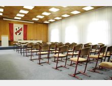 Konferenzraum mit einer Bestuhlung aus gepolsterten Stühlen in Reihen, einem Rednerpult und einem Tisch auf einer erhöhten Bühne. Die Wände sind aus Holz, mit einem großen Wappenbanner dekoriert, und seitlich befinden sich große Fenster mit weißen Vorhängen.