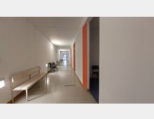 Langer, schlichter Flur in einem Gebäude mit weißen Wänden, vereinzelten orangefarbenen Türrahmen und mehreren Wartebänken an den Seiten. Der Boden ist beige, und am Ende des Flurs befindet sich eine verglaste Tür mit Blick nach draußen.