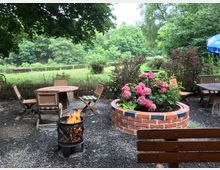 Ein Gartenbereich mit Kiesboden, umgeben von grünen Bäumen und Wiesen. Im Vordergrund stehen Holztische und -stühle, eine Feuerstelle mit Flammen sowie ein rundes Hochbeet aus Ziegelsteinen mit rosa Hortensien.
