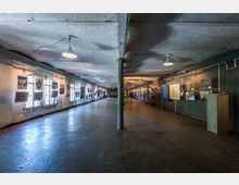 Langer Ausstellungsraum in einem industriellen Gebäude mit Betonboden, weißen Wänden und freiliegender Decke. Links befinden sich Fenster mit aufgehängten Bildern, rechts sind verschiedene Informationstafeln und Exponate an der Wand befestigt.