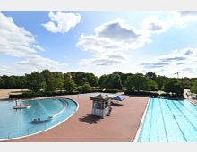 Ein Freibad mit zwei Becken, eines in geschwungener Form mit Wasserspielen und eines rechteckig für Schwimmbahnen. Umgeben von Bäumen und Parkflächen, mit einem roten Pflasterboden und einer kleinen Überdachung zwischen den Becken.