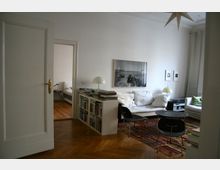 Heller Wohnraum mit Parkettboden, weißem Sofa, Couchtisch und buntem Teppich, dazu ein Bücherregal als Raumteiler und mehrere Tischlampen. An der Wand hängen gerahmte Fotos, und durch eine offene Tür ist ein angrenzendes Schlafzimmer zu sehen.