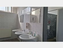 Gefliester Waschraum mit drei wandmontierten Waschbecken, Spiegeln und Seifenspendern, beleuchtet durch ein hohes Fensterband. Rechts ist ein angrenzender Bereich mit grauer Wand und sichtbaren Kabinenabtrennungen zu erkennen.