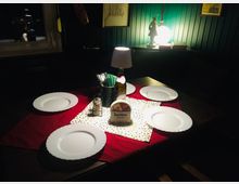 Ein Tisch in einem gemütlichen Restaurant-interieur, gedeckt mit vier weißen Tellern auf einer roten Tischdecke, flankiert von Servietten, einer Tischlampe und Dekorationsgegenständen wie einer Flasche und einem Utensilienhalter. Im Hintergrund ist eine dunkelgrüne Wand mit Bildern und Beleuchtung zu sehen.