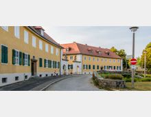 Gelbe historische Gebäude mit roten Ziegeldächern und grünen Fensterläden an einer gepflasterten Straße. Ein Torbogen mit dekorativen Statuen verbindet zwei Gebäudeteile, und im Vordergrund sind ein Steinbeet sowie Straßenschilder sichtbar.
