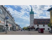 Weitläufiger Platz in einer Innenstadt mit einem weißen Rathausgebäude, das einen hohen Uhrturm mit grünem Spitzdach besitzt. Links befinden sich moderne Geschäftshäuser mit Schaufenstern und Schildern, rechts ist ein Teil eines Gebäudes mit Satteldach und Rundfenstern zu sehen.