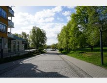 Pflastersteinstraße mit angrenzendem Gehweg führt zu einem Flussufer, umgeben von grünen Bäumen. Links steht ein modernes Gebäude mit Balkonen, während rechts eine Rasenfläche mit Parkbeleuchtung zu sehen ist.