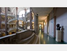 Innenansicht eines historischen Gebäudes mit gewundenen, dekorativen Treppen und verzierten Geländern. Der Raum ist geprägt von hohen Decken mit Stuckmustern, großen Fenstern, Säulen und einer seitlich stehenden Büste auf einem Sockel im Vordergrund.