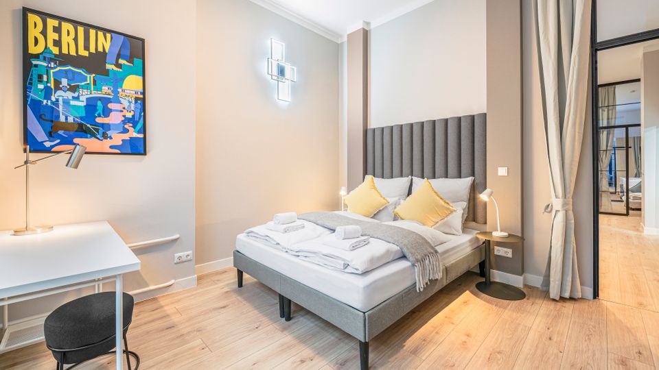 Modernes Schlafzimmer mit Doppelbett, hohem gepolstertem Kopfteil, Nachttischlampen und hellem Holzboden. Links steht ein weißer Schreibtisch unter einem „Berlin“-Poster, rechts trennt eine Glaswand mit Vorhang den Raum zu einem Flur oder Nebenbereich.