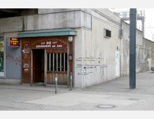 Außenansicht einer kleinen Bar an einer Hausecke mit dem Namen „Besenkammer-Bar – Die Szenekneipe am Alex“. Die Fassade besteht aus Holz und Beton, mit Schriftzügen und einem kleinen Vordach. Rechts daneben eine schlichte Betonwand mit Graffiti und Linienzeichnungen.