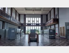 Innenansicht eines historischen Kraftwerkskontrollraums mit hohen Decken, großen Fenstern und einem umlaufenden Balkon. Im Raum stehen metallene Schalttafeln, und in der Mitte befindet sich eine Treppe, die nach unten führt.