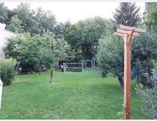 Großer, gepflegter Garten mit Rasenfläche, Obstbäumen und dichtem Baumbestand am Rand; in der Mitte steht ein kleines Fußballtor. Rechts im Vordergrund befindet sich ein Holzpfosten mit Querbalken, daneben Sträucher und ein Zaun.