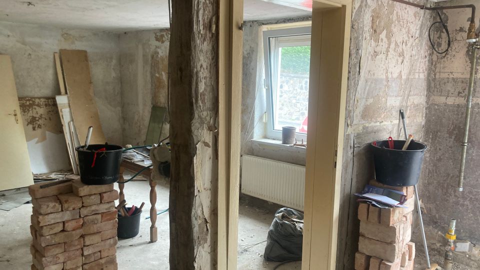 Ein renovierungsbedürftiger Innenraum mit unverputzten Wänden, einem offenen Türrahmen und sichtbarem Mauerwerk. Im Raum stehen gestapelte Ziegelsteine, Eimer mit Werkzeugen und Baumaterialien, sowie ein Fenster, das Tageslicht hereinlässt.