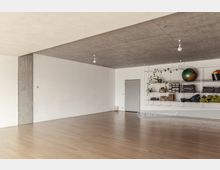 Ein minimalistischer Raum mit Holzboden, weiß gestrichenen Wänden und einer Sichtbetondecke. An einer Wand befindet sich ein offenes Regal mit Yogamatten, Handtüchern, einer Pflanze und verschiedenen Gegenständen; daneben eine geschlossene Tür.