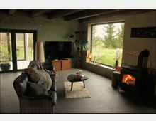 Wohnzimmer in einem rustikalen Stil mit sichtbaren Holzbalken und verputzten Wänden. Im Raum befinden sich ein Kaminofen, eine Ledercouch, ein kleiner Couchtisch auf einem Teppich, sowie ein großes Fenster mit Blick auf eine grüne Landschaft.