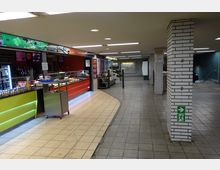 Ein U-Bahnhof mit gastronomischen Angeboten, darunter ein Imbiss mit leuchtend farbiger Theke sowie einer Auslage mit Backwaren. Die Umgebung ist mit hellen Fliesen, weißen Kachelpfeilern und einer Notausgangsbeschilderung gestaltet, im Hintergrund stehen Blumenstände und ein Fahrkartenautomat.