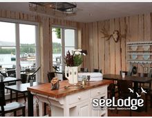 Innenansicht eines gemütlichen Restaurantbereichs mit Holzwänden und rustikaler Dekoration, darunter Geweihe und ein Regal mit Tellern. Im Vordergrund steht ein Holztresen mit Blumen und dekorativen Gegenständen, während große Fenster den Blick auf eine Terrasse und einen See mit Booten freigeben.