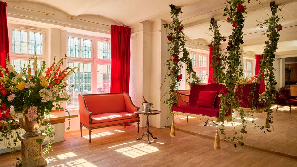 Heller Salonraum mit großen Sprossenfenstern und roten Vorhängen, Holzboden sowie Sitzmöbeln in Rot. In der Mitte hängt eine dekorative Schaukelbank, umrankt von grünen Ranken mit roten Rosen; daneben stehen ein kleiner Beistelltisch und ein großer Blumenstrauß auf einer Steinsäule.