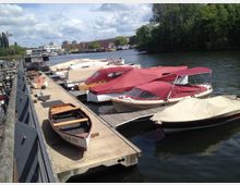Bootsanlegestelle an einem Fluss mit mehreren Anlegeplätzen, auf denen motorisierte und hölzerne Boote mit Abdeckplanen festgemacht sind. Im Hintergrund sind grüne Bäume, einige Gebäude und ein teils bewölkter Himmel zu sehen.