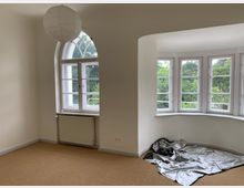 Ein leerer Raum mit hellen Wänden, einem großen, rundbogigen Fenster auf der linken Seite und einem dreiteiligen Erkerfenster rechts mit Blick ins Grüne. Der Boden ist mit Teppich ausgelegt, eine papierne Hängelampe und ein Heizkörper befinden sich unter dem linken Fenster, während im Erkerbereich eine Plastikplane liegt.